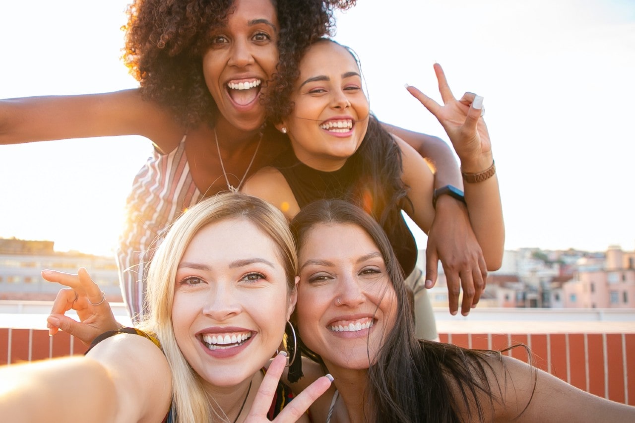 Four women smile joyfully, taking a selfie outdoors with a cityscape backdrop.