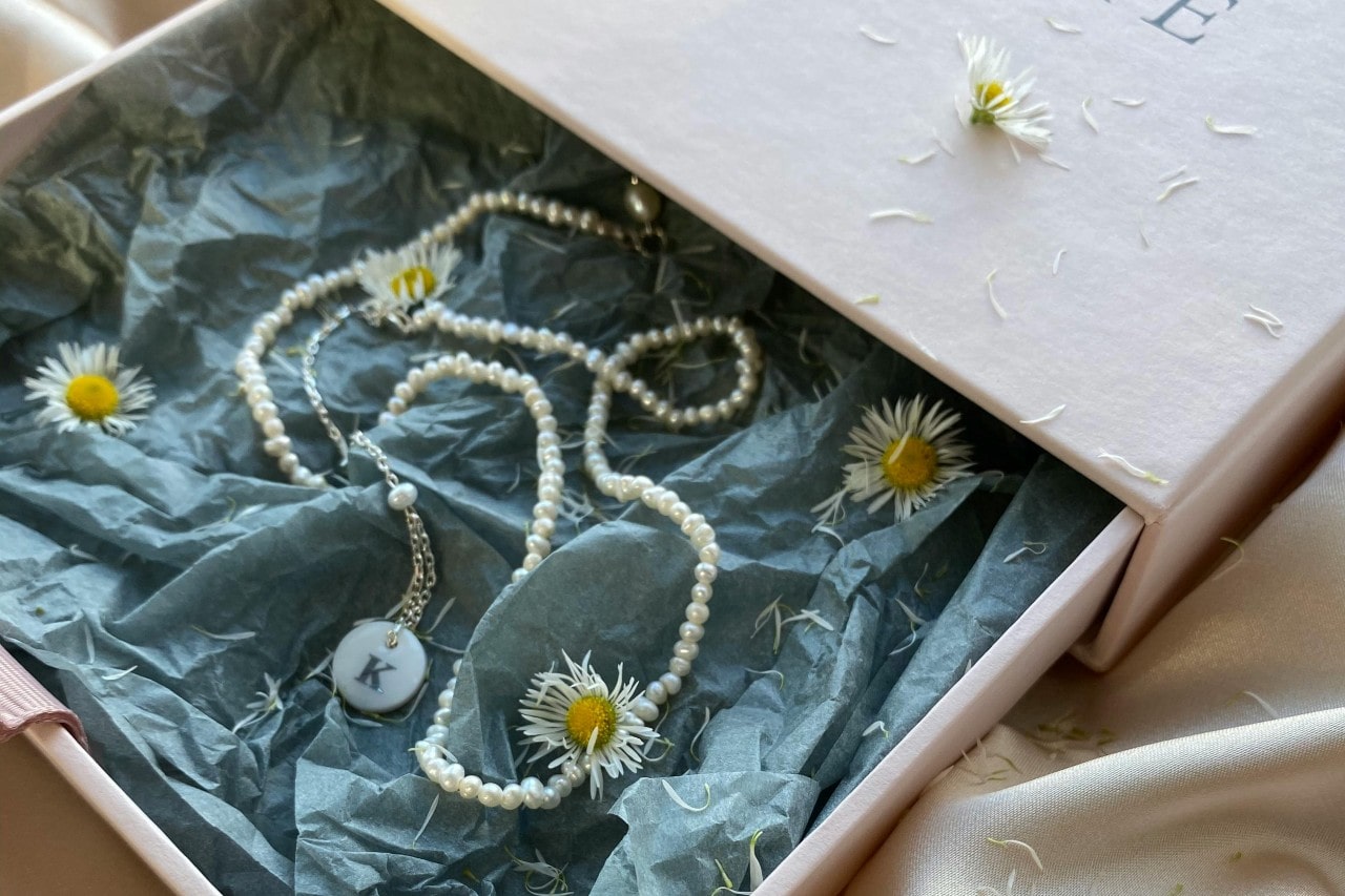 A delicate pearl necklace with a pendant rests in a pastel pink box lined with blue tissue paper, adorned with daisies.