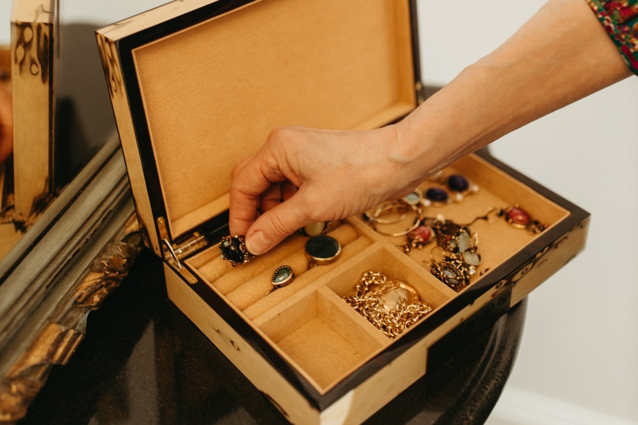 Hand reaching into an open jewelry box with assorted vintage rings, necklaces, and earrings on a dark wooden surface.