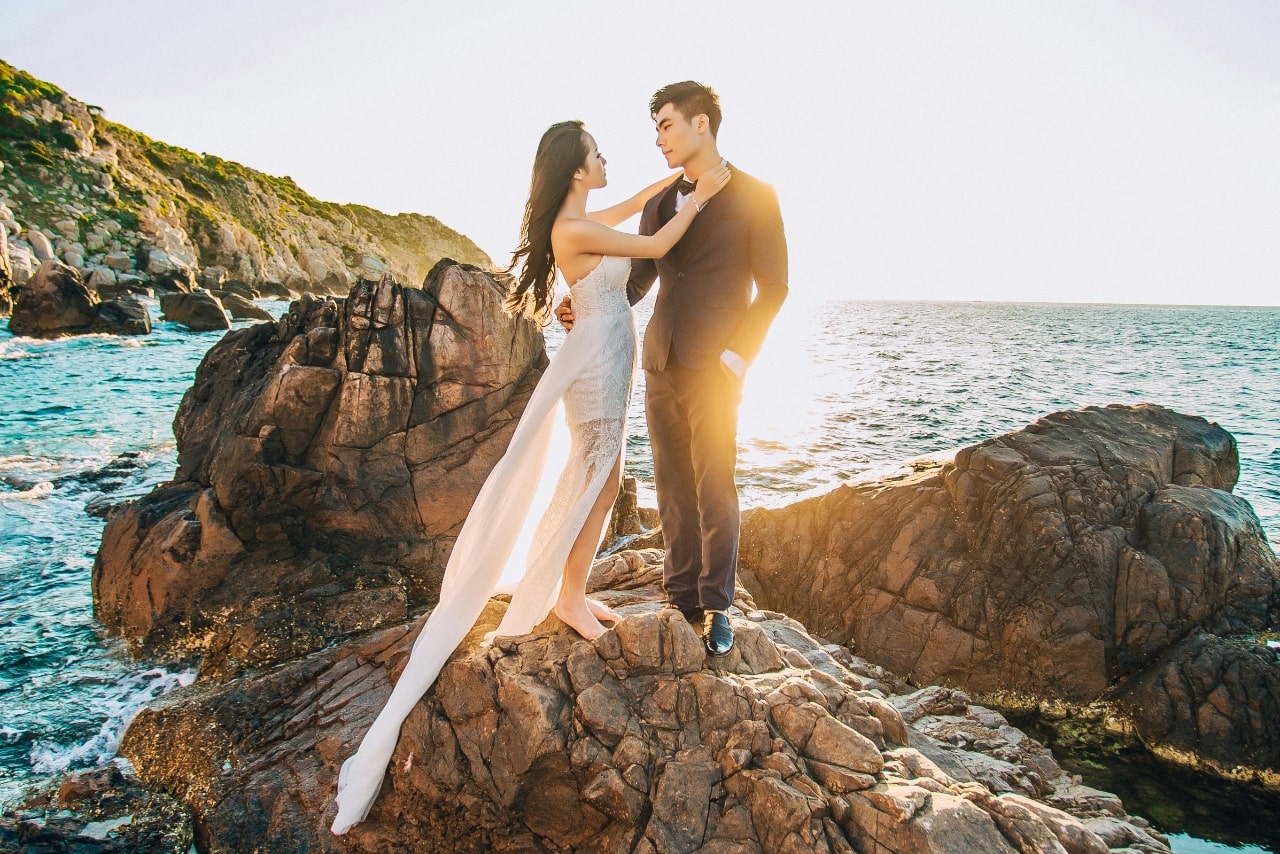 A wide view of a newlywed couple standing on beach rocks on a beautiful, sunny day.