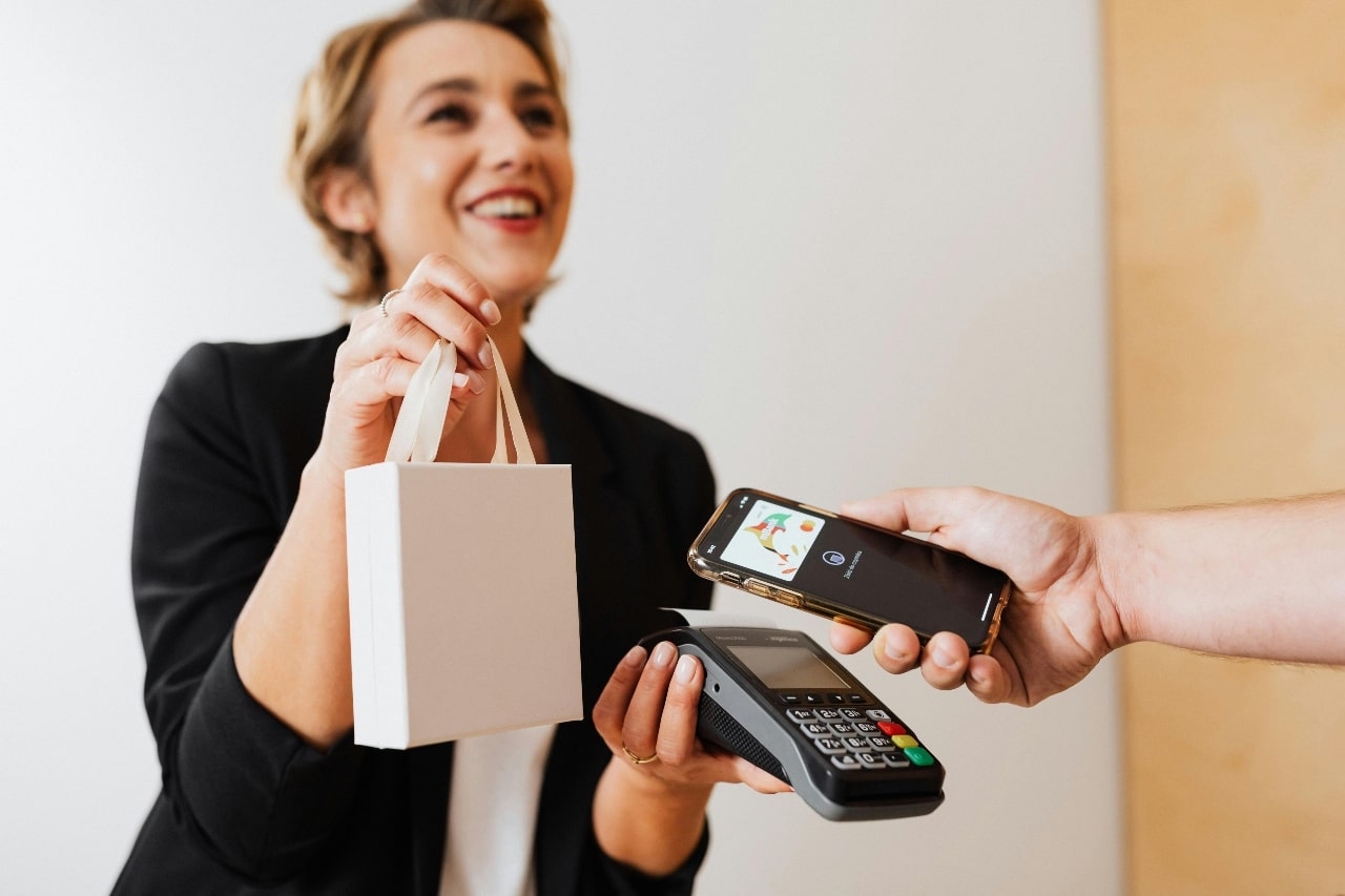 A smiling cashier holds a payment terminal and a shopping bag while a customer completes a purchase.