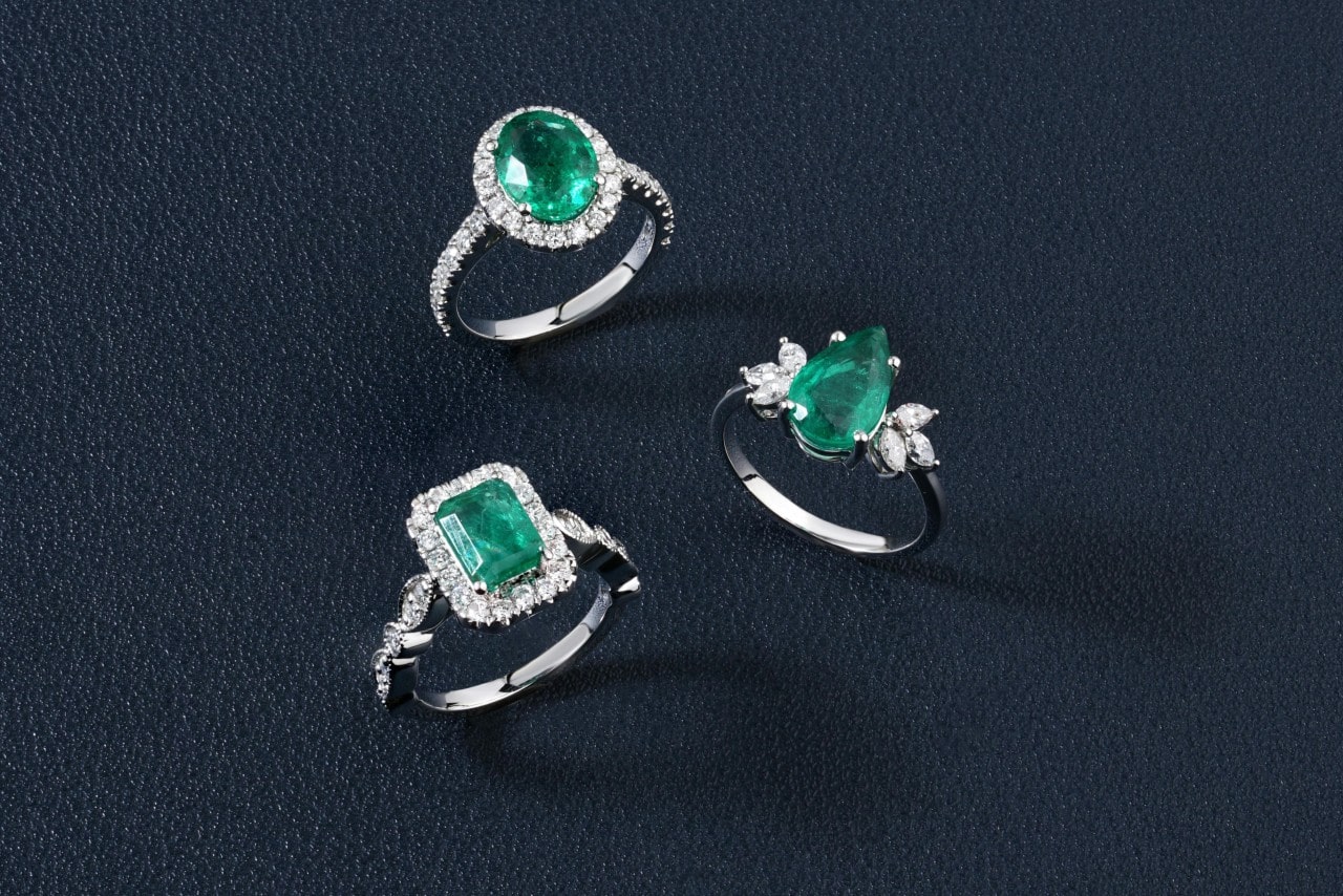 Three elegant rings with vibrant green emeralds, adorned with sparkling diamonds, displayed on a dark textured surface