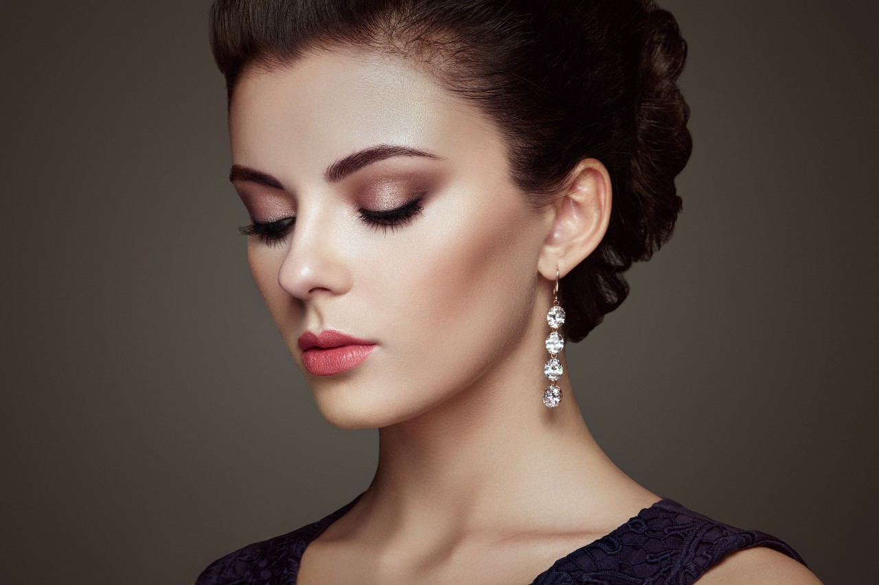A woman with makeup and an updo hairstyle wears a long, sparkling drop earring with multiple round diamonds.