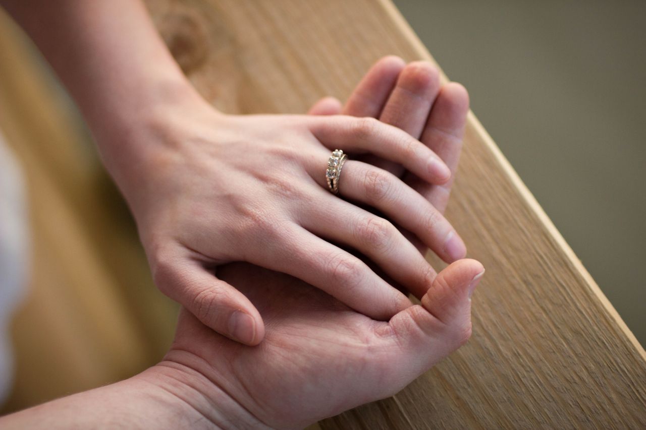 A close-up of a couple holding hands on a wooden rail, with emphasis on the engagement ring on her finger.