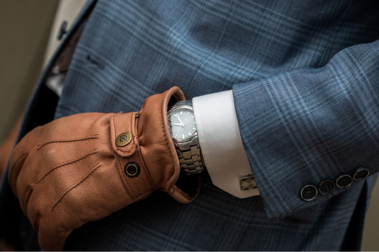 A person in a blue checkered suit wearing a luxury silver watch partially covered by a brown leather glove.