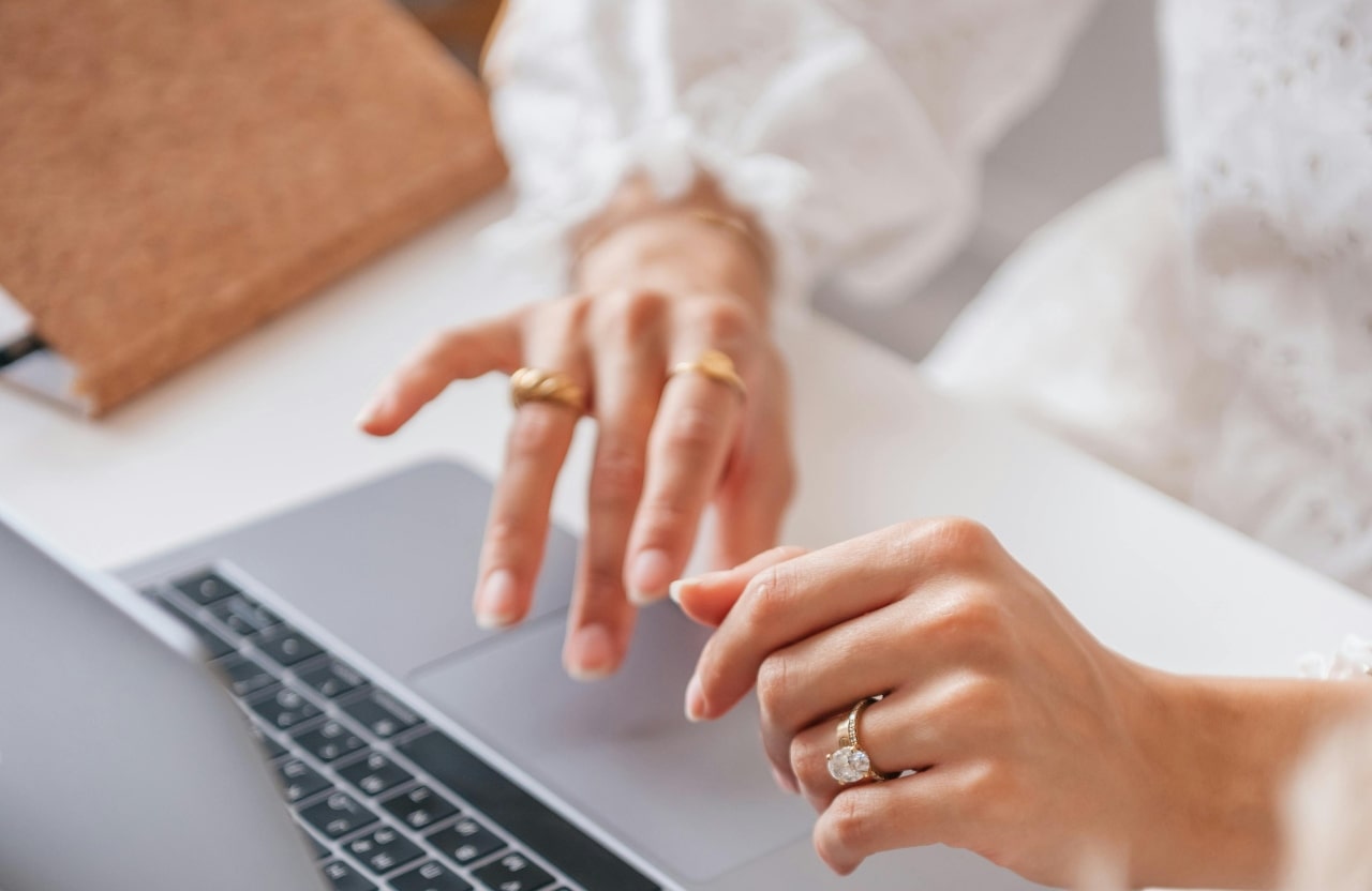 A close-up of a woman’s hands typing on a laptop, adorned with multiple rings, including an engagement ring on her left hand.