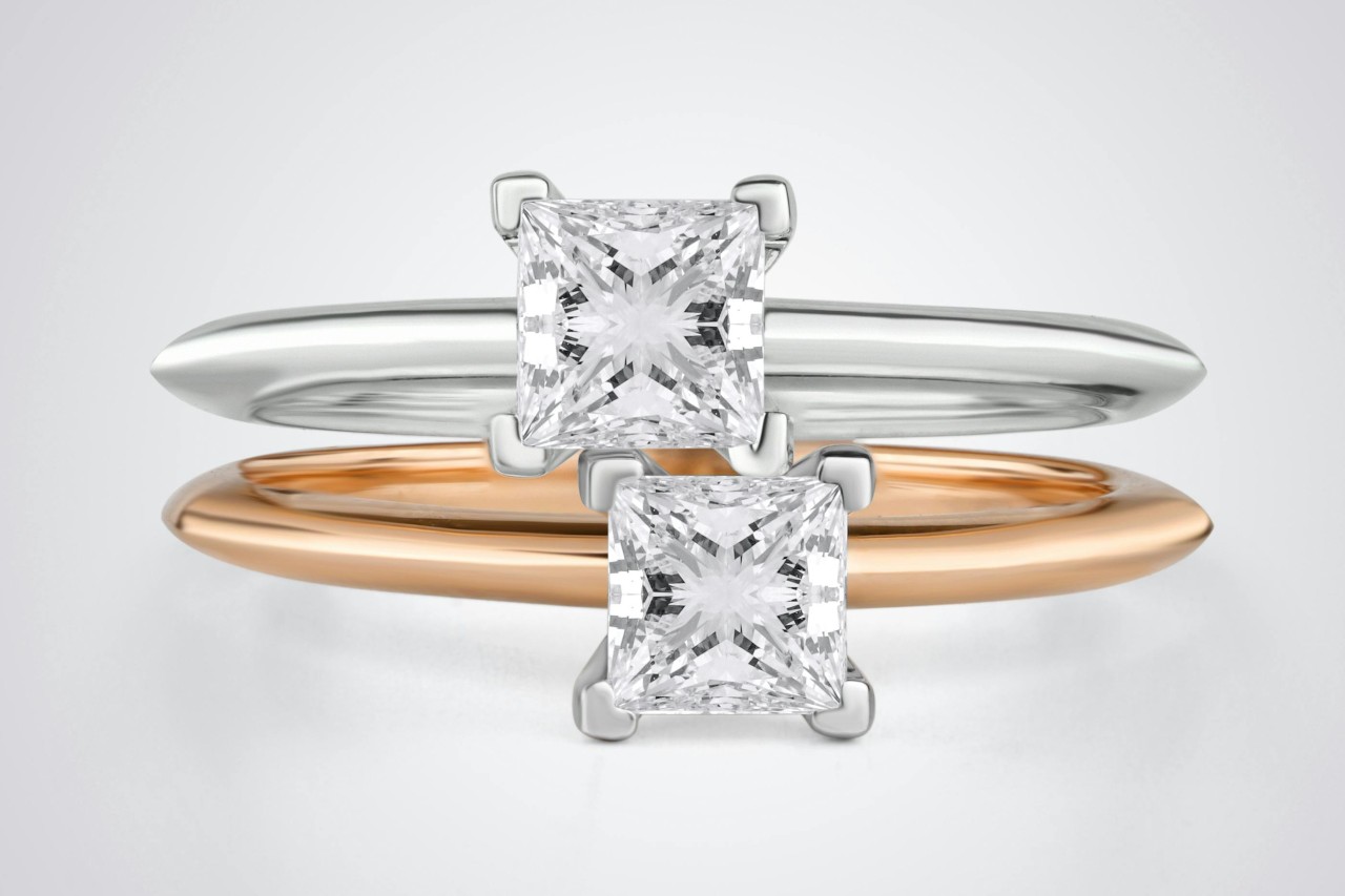 A close-up of two princess cut solitaire engagement rings, one crafted in platinum and the other in rose gold.