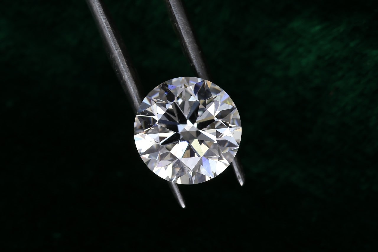 A close-up of a pristine round cut diamond held in a pair of jeweler’s tweezers on a dark green backdrop.
