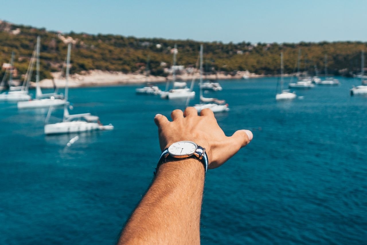 A man’s hand adorned with a blue and white strapped watch in front of the sea full of yachts.