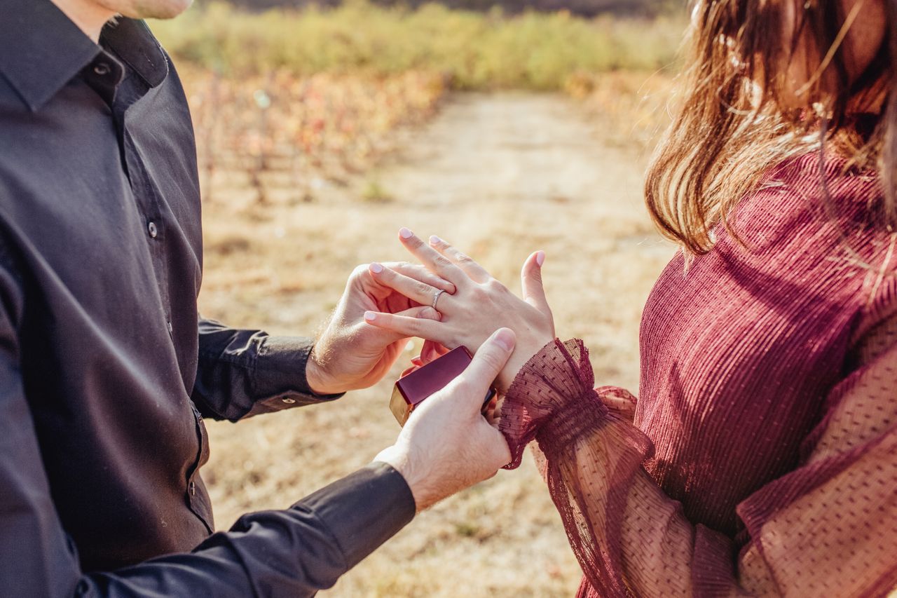 A man slips a platinum engagement ring on his lover’s finger while outside.