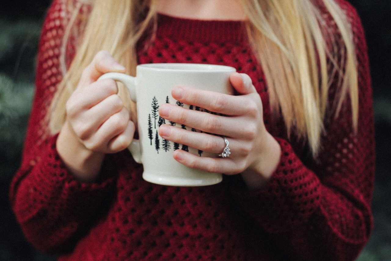A woman wearing a sweater sips coffee while wearing her contemporary white gold engagement ring.