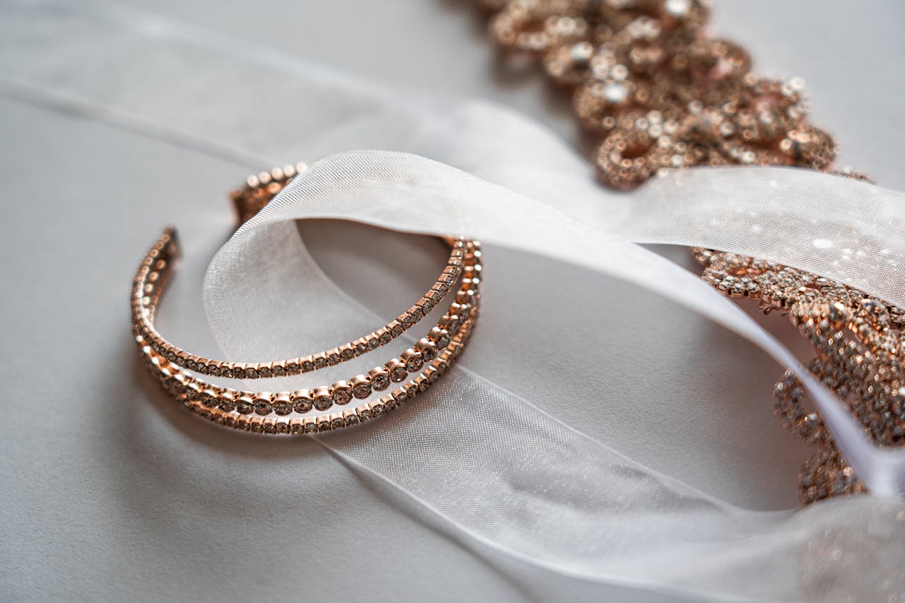 A close-up of an elegant rose gold bracelet intertwined with a sheer white ribbon against a soft white background.