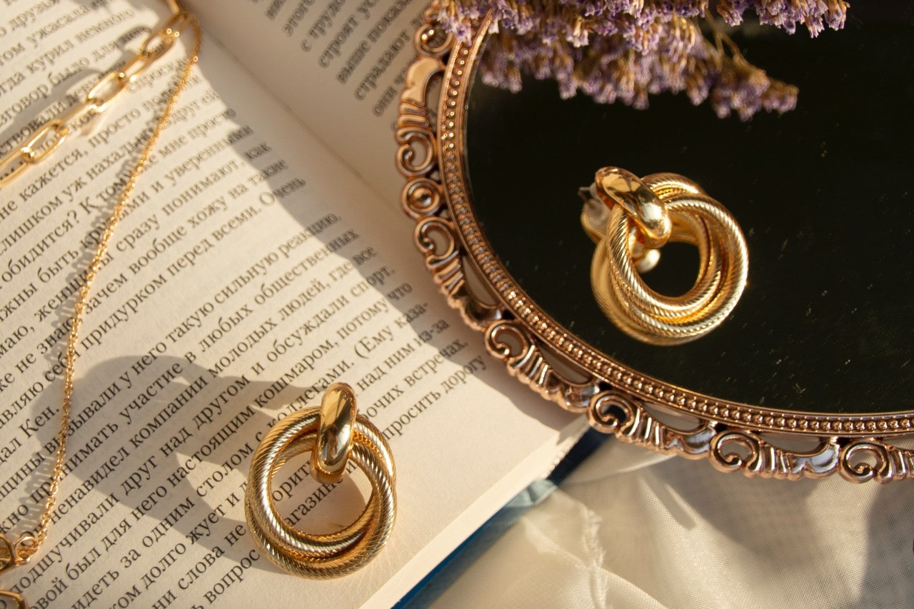 A set of gold intertwined hoop earrings placed on an open book next to a mirror and delicate gold jewelry. A set of gold intertwined hoop earrings placed on an open book next to a mirror and delicate gold jewelry.