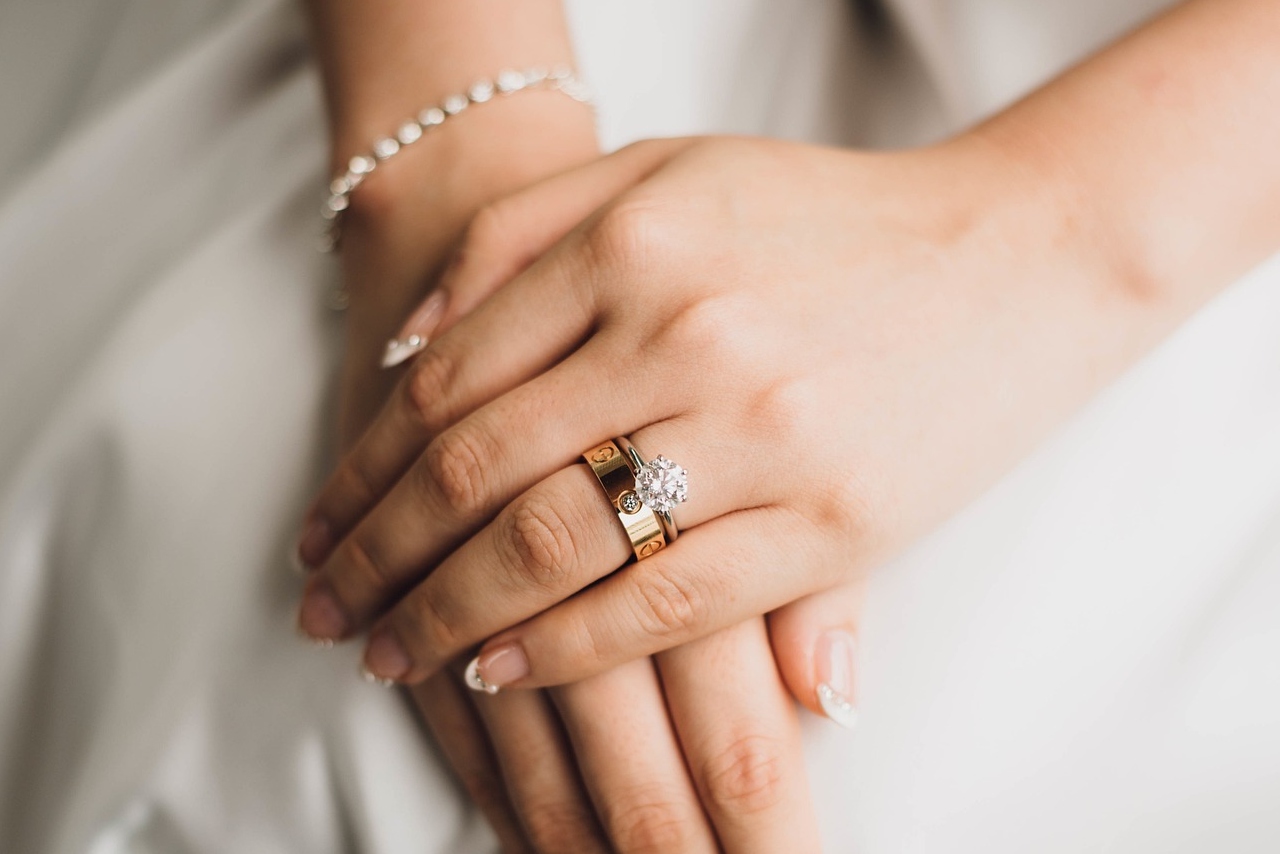 A close-up of a woman’s hand wearing a white gold engagement ring with a stacked yellow gold band, resting on her lap in a white dress