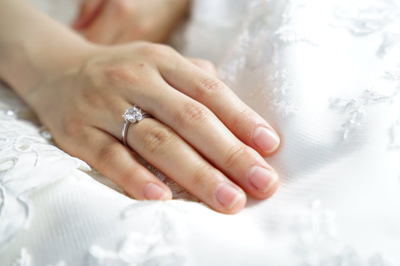 A close up of a bride’s hand with a solitaire engagement ring glistening on the ring finger, resting on a white wedding gown.