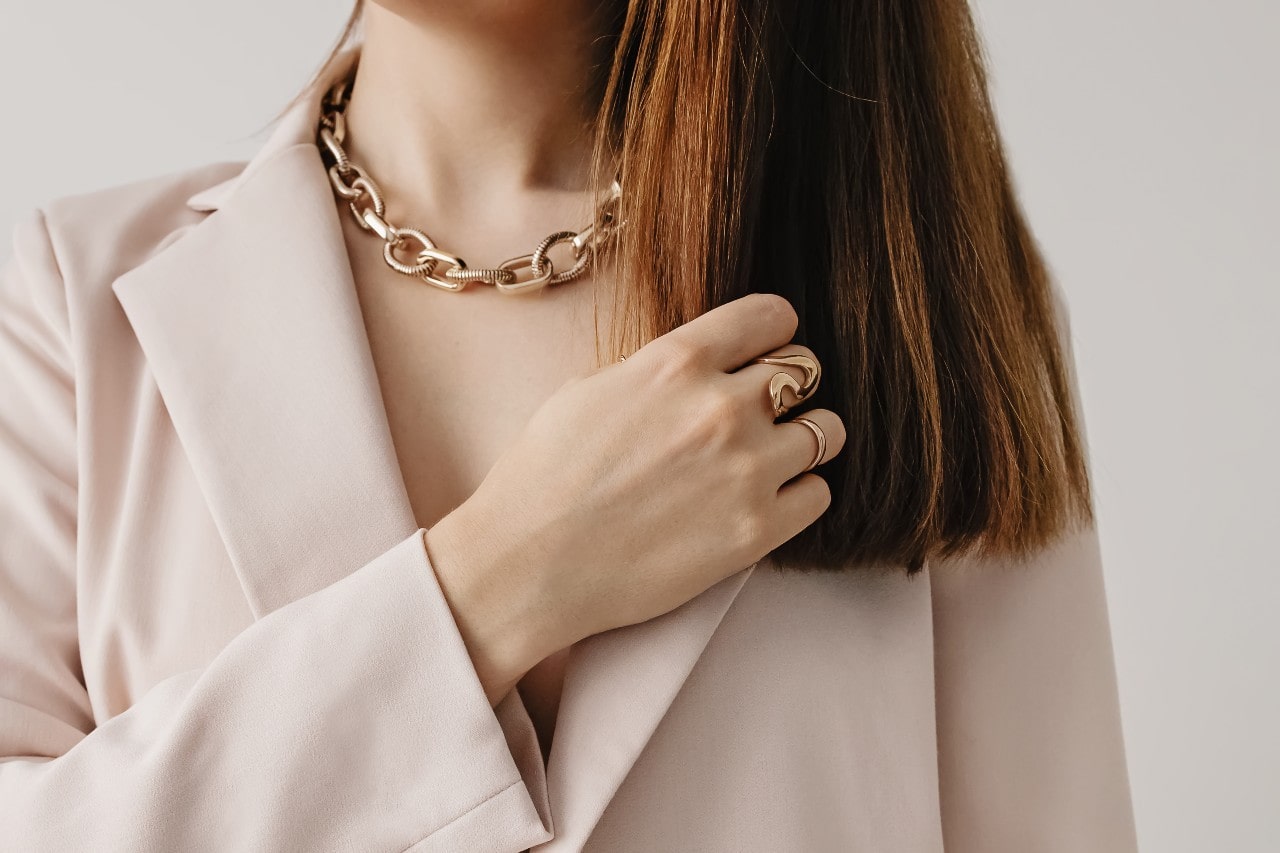 A woman in a beige blazer showcases a bold yellow gold chain necklace and statement rings, with her hand touching her hair. A woman in a beige blazer showcases a bold yellow gold chain necklace and statement rings, with her hand touching her hair.