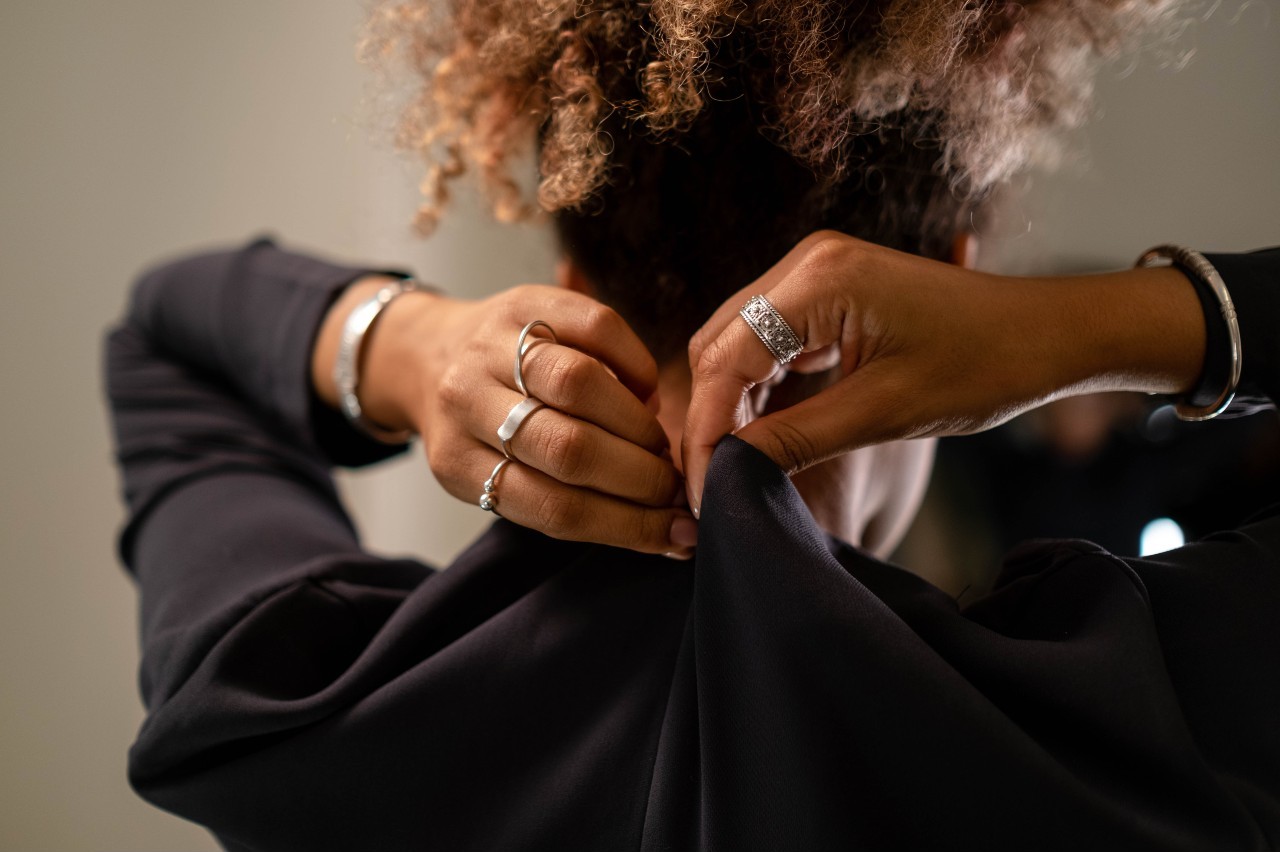A woman with curly hair is seen from behind, fastening a button on a black garment, adorned with silver rings and bracelets on both hands. A woman with curly hair is seen from behind, fastening a button on a black garment, adorned with silver rings and bracelets on both hands.