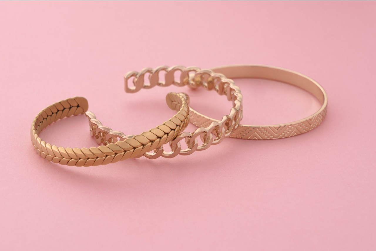 Three yellow gold open bracelets with different textures and patterns on a pink surface. Three yellow gold open bracelets with different textures and patterns on a pink surface.