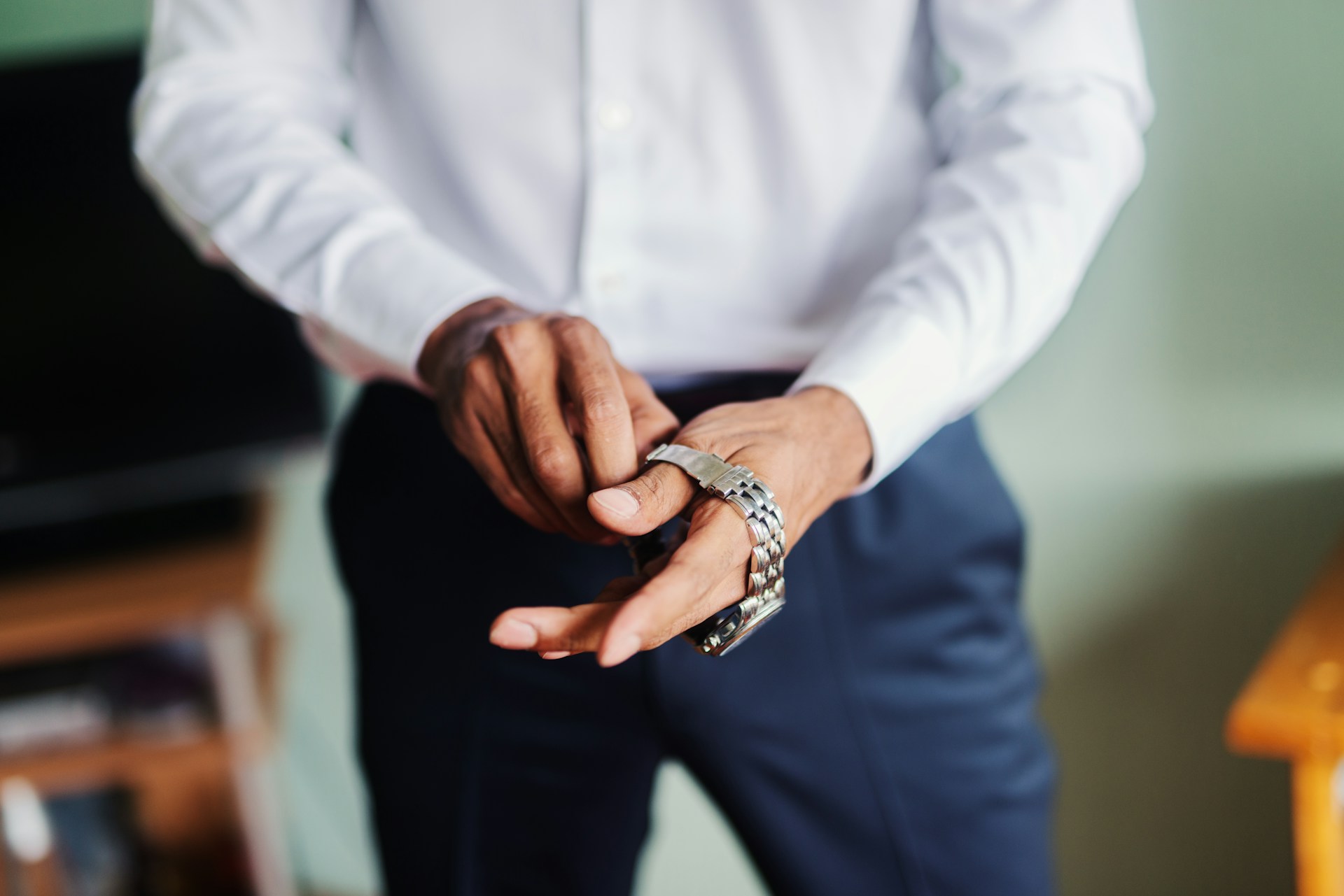 A close-up of a man in a white shirt and blue pants putting on a stainless steel watch.