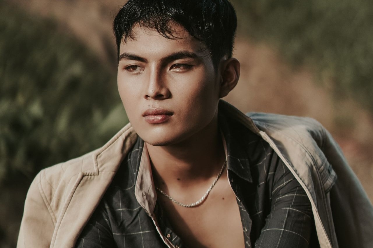 A man wearing a chain necklace and two layers of shirts, while sitting against a blurry landspace A man wearing a chain necklace and two layers of shirts, while sitting against a blurry landspace