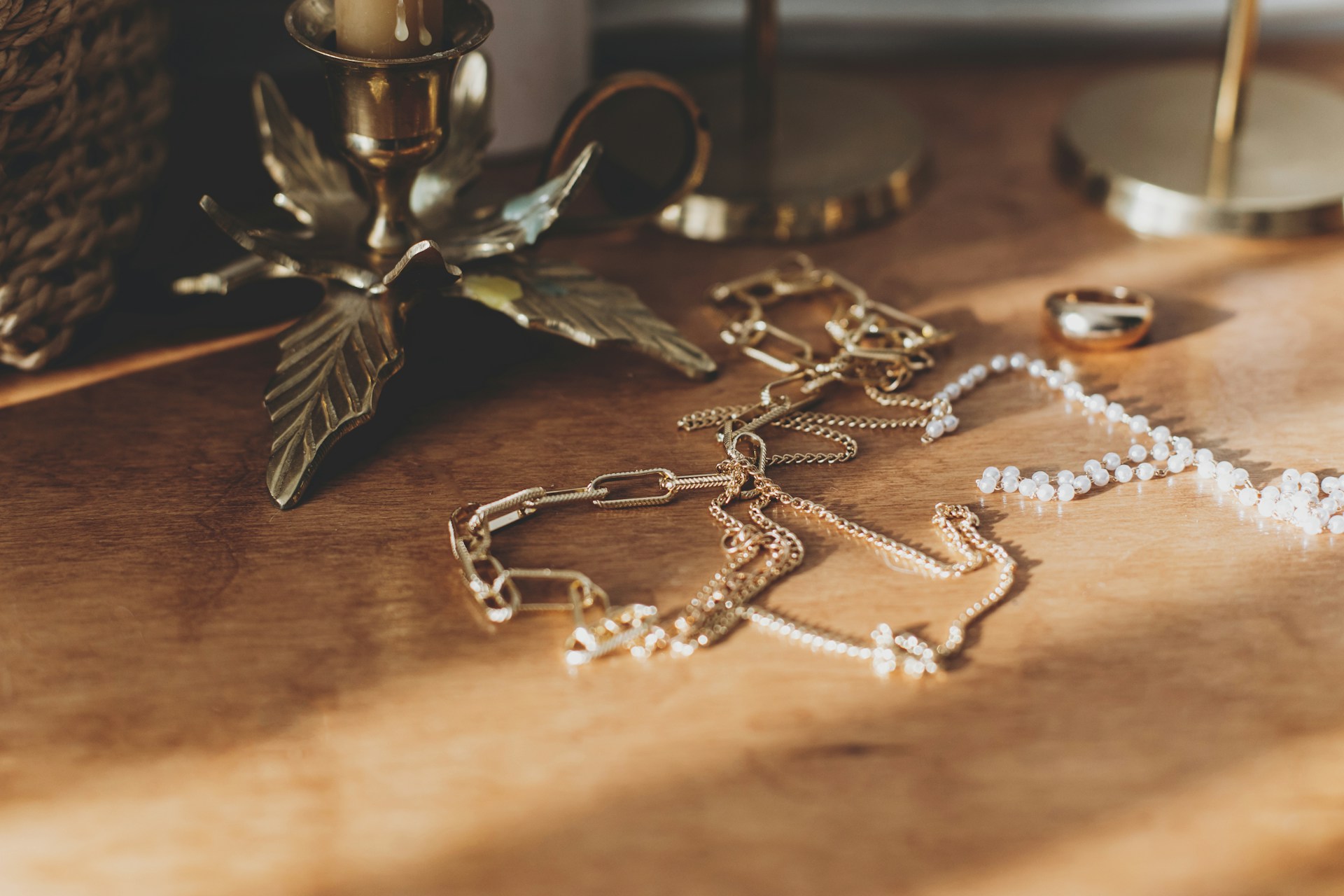 A variety of gold chains, pearl necklaces, and a ring are laid out on a wooden surface beside brass candle holders. A variety of gold chains, pearl necklaces, and a ring are laid out on a wooden surface beside brass candle holders.
