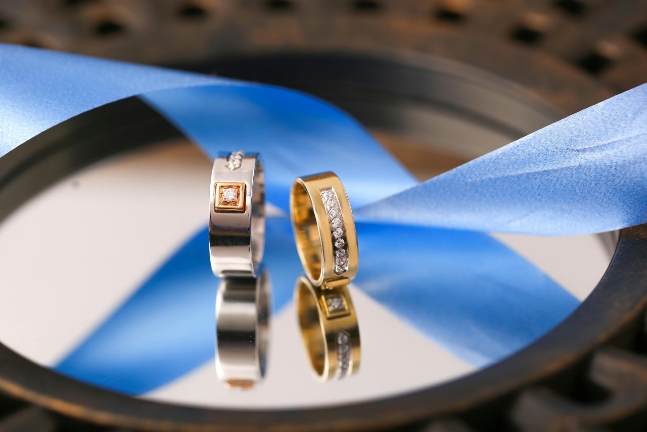 White and yellow gold diamond wedding bands on a reflective surface near the blue ribbon.