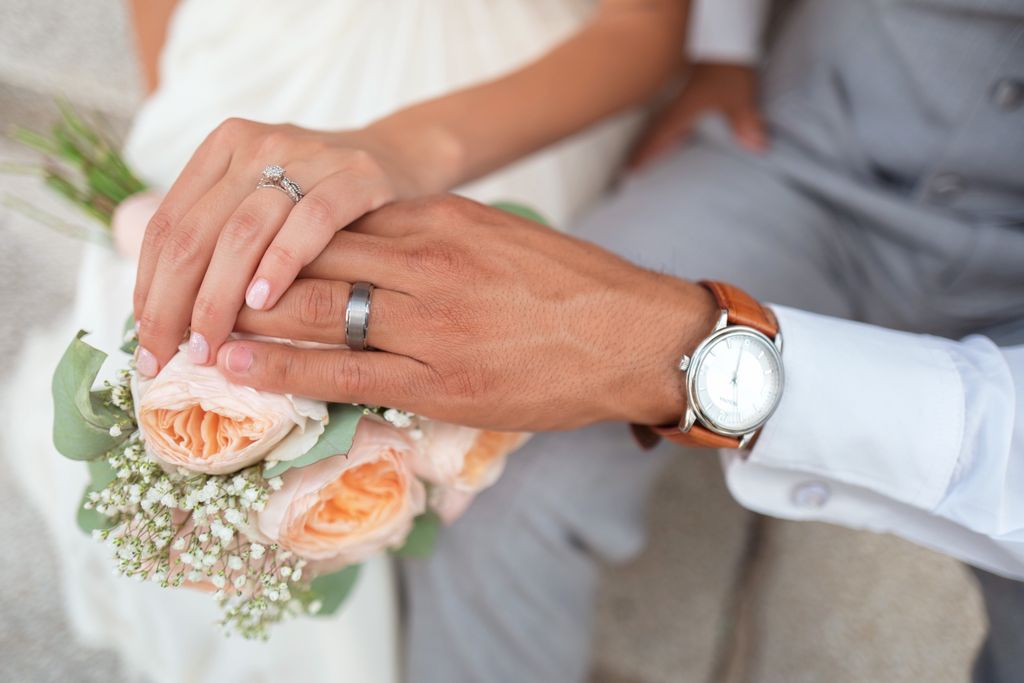 A close-up of a bride and groom with their hands atop a bouquet of roses, with emphasis on their wedding bands and the groom’s wristwatch.
