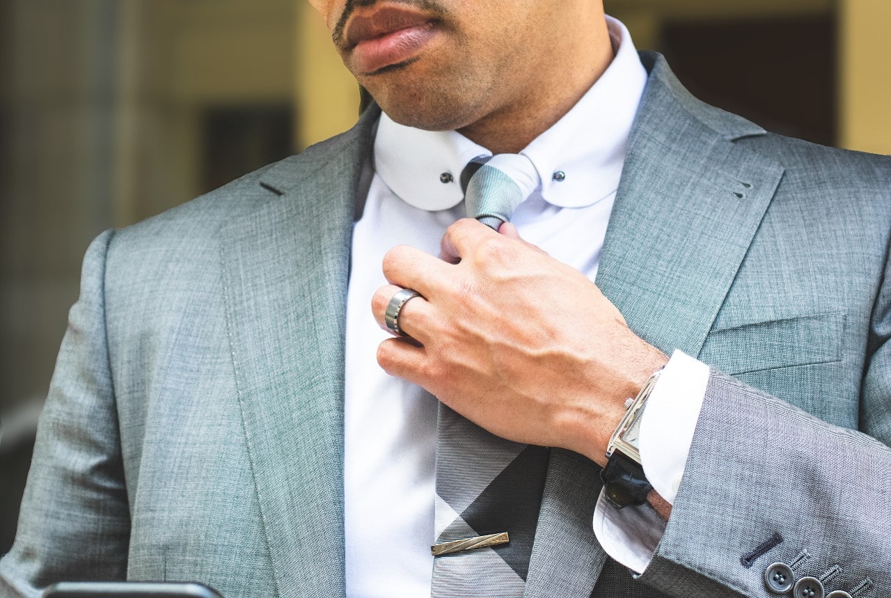 A close-up of a man in a grey-blue suit fixing his tie, with emphasis on his bold silver-toned wedding band.