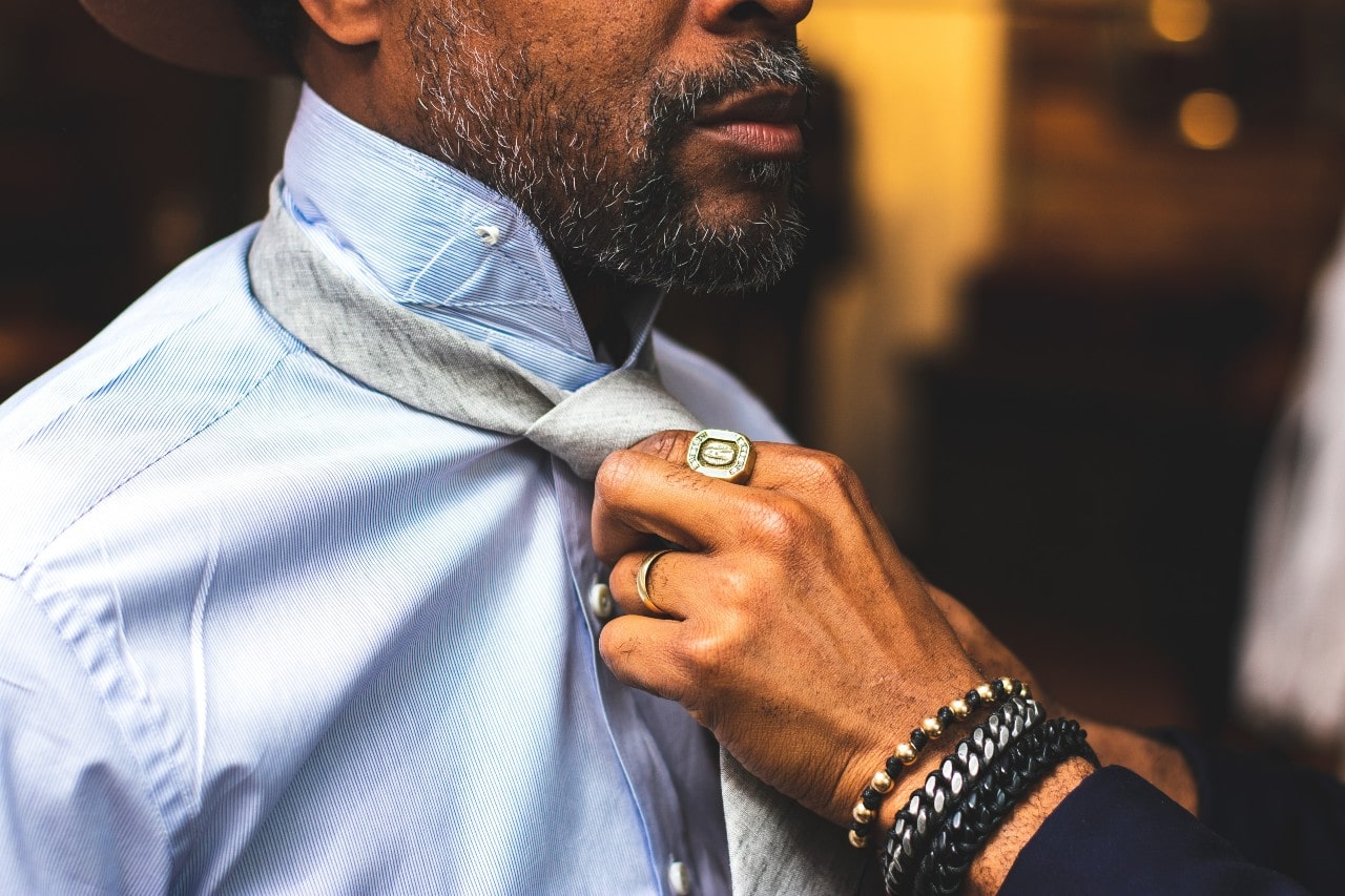 A close up of a man’s hand, clad in a yellow gold wedding band, a distinctive signet ring, and multiple chain bracelets, tying another man’s tie.