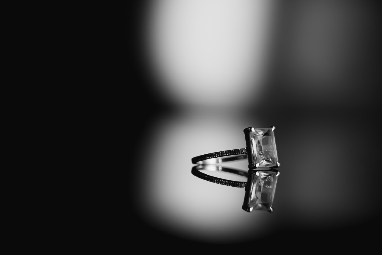 A solitaire ring with an emerald-cut diamond sits on a reflective surface.