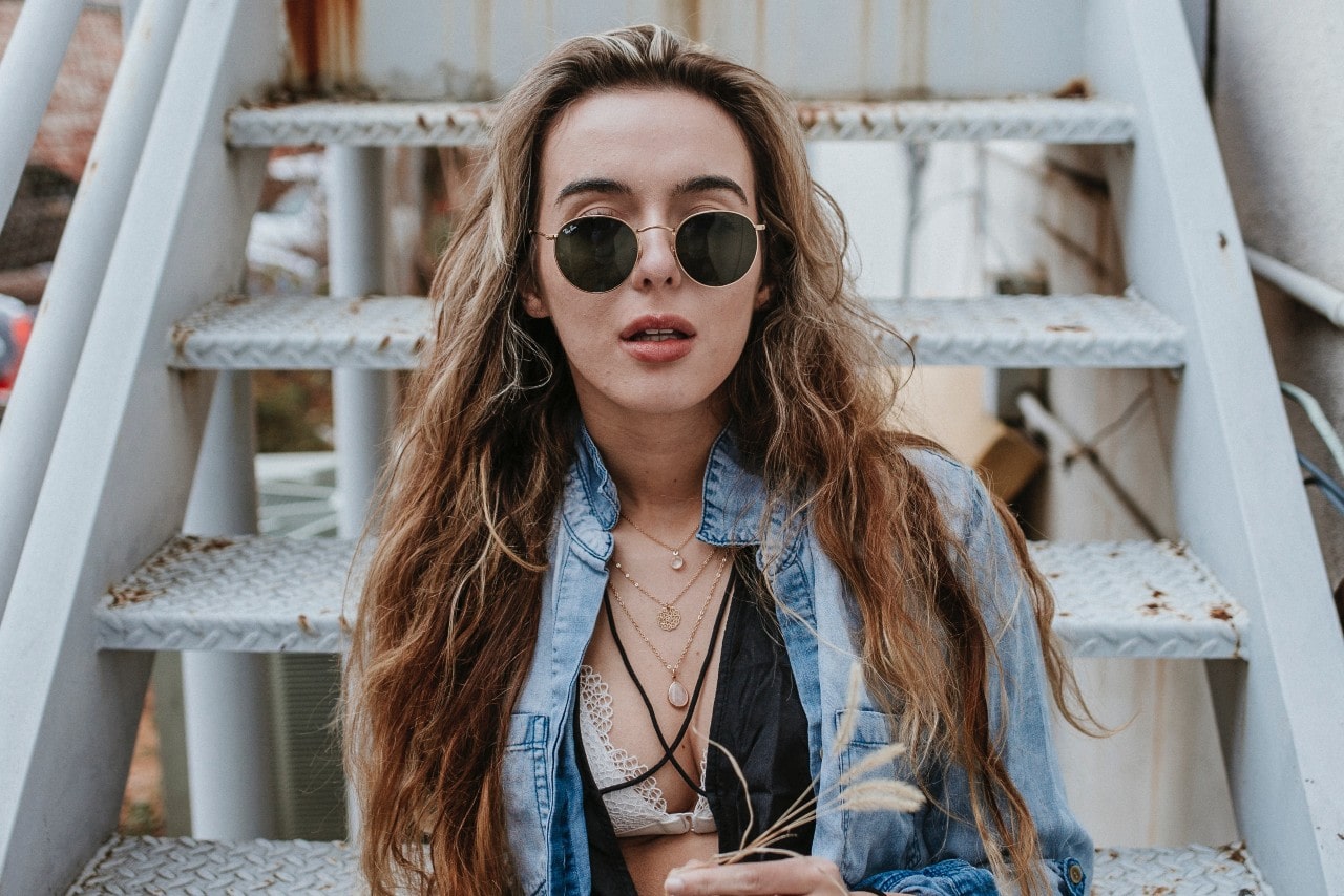A blond woman with sunglasses and denim jacket sits on metallic stairs, showcasing a layered gold chain necklace.