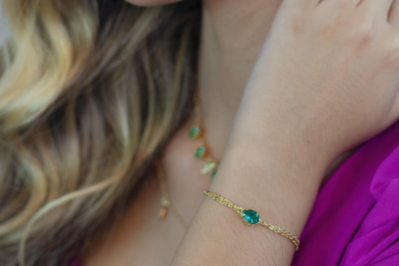 A close up of the woman's wrist and neck, showcasing a yellow gold gemstone bracelet and necklace.