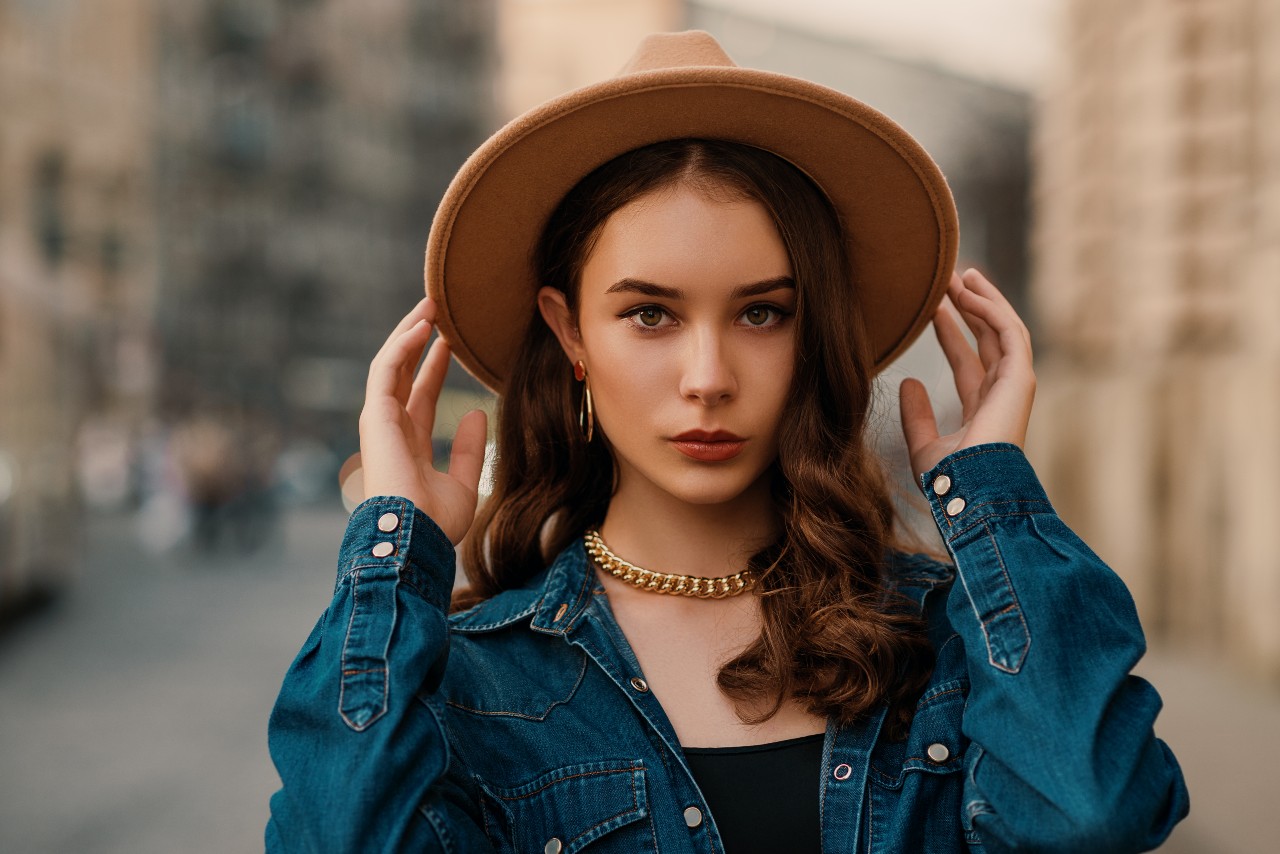 A young woman in a denim shirt and a brown hat, wearing a chunky yellow gold chain necklace and hoop earrings.