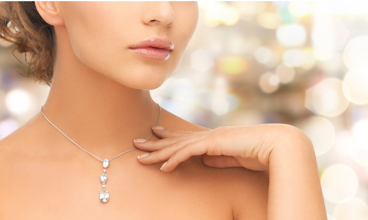 A close up of a woman wearing a white gold necklace with three vertically aligned diamonds, shown against a soft bokeh background.