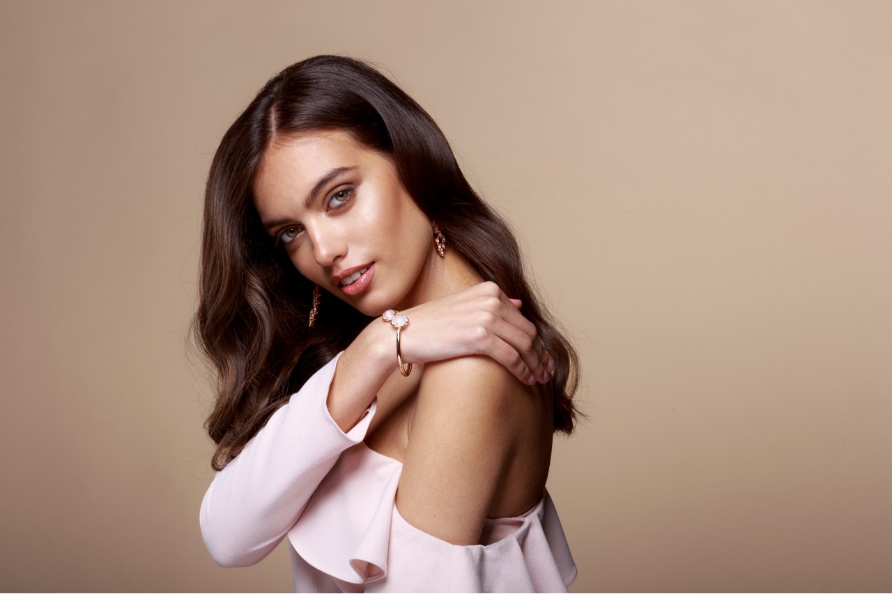 A woman with wavy brown hair and a pink strapless top showcases rose gold floral bangle and drop earrings.