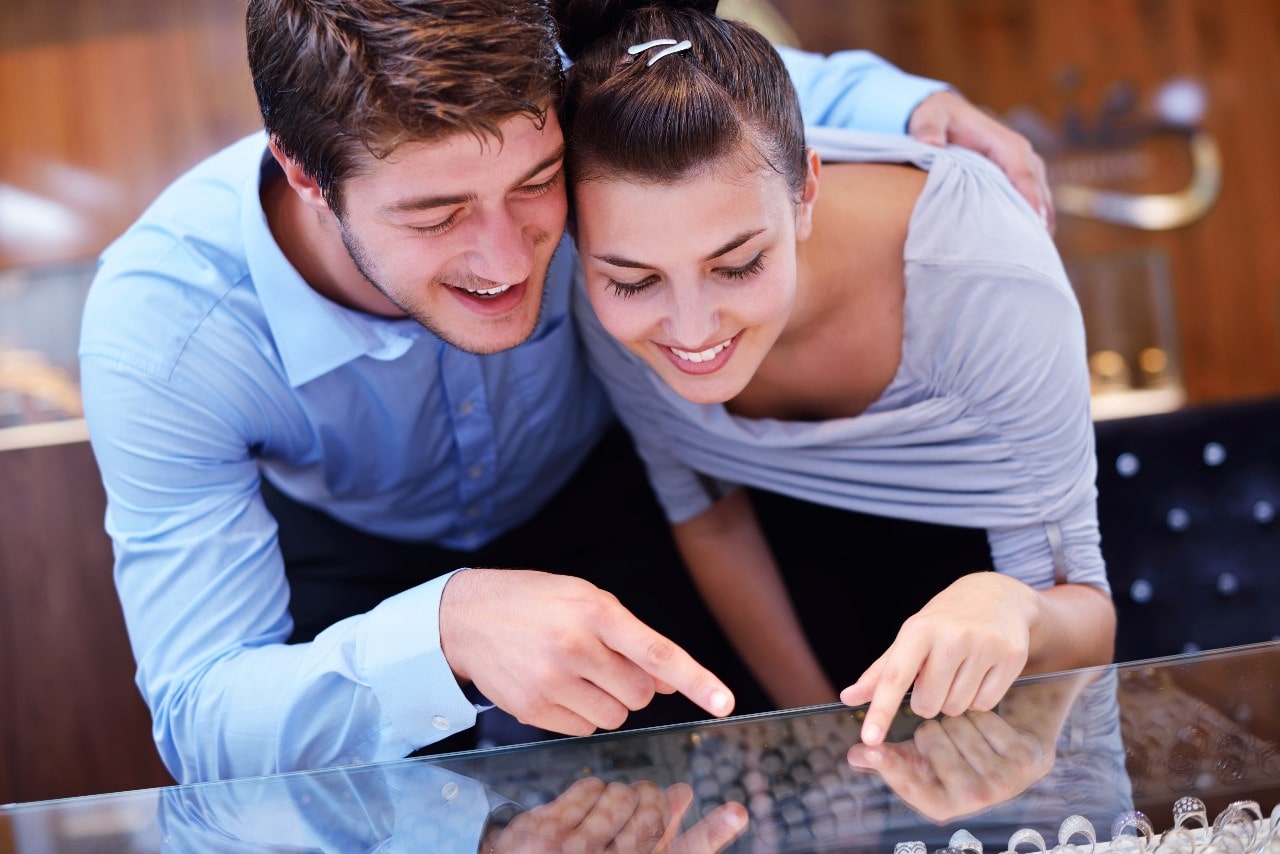 A smiling couple in casual attire lean over a jewellery display case, pointing at rings.