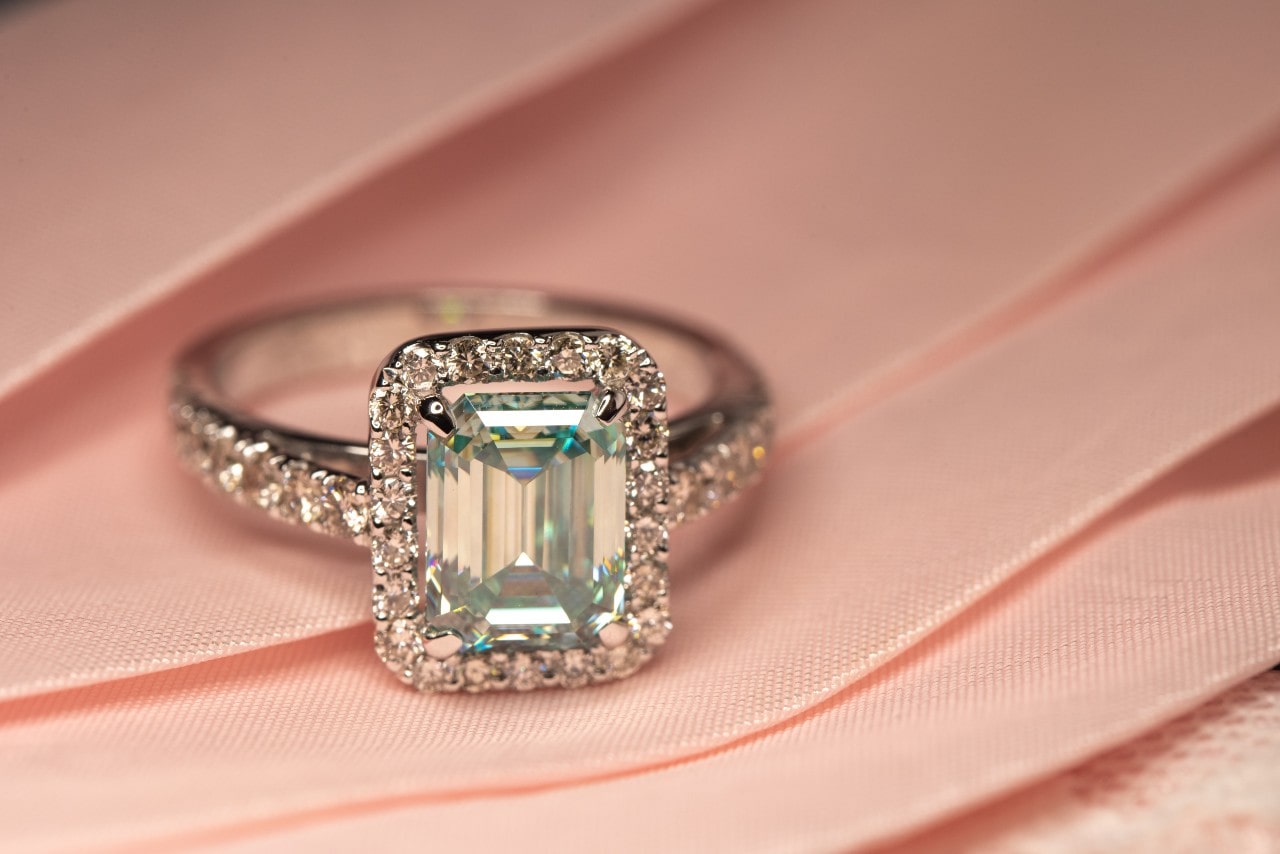 A radiant diamond ring with a rectangular emerald-cut stone, surrounded by smaller diamonds on a silver band, rests on soft pink fabric.