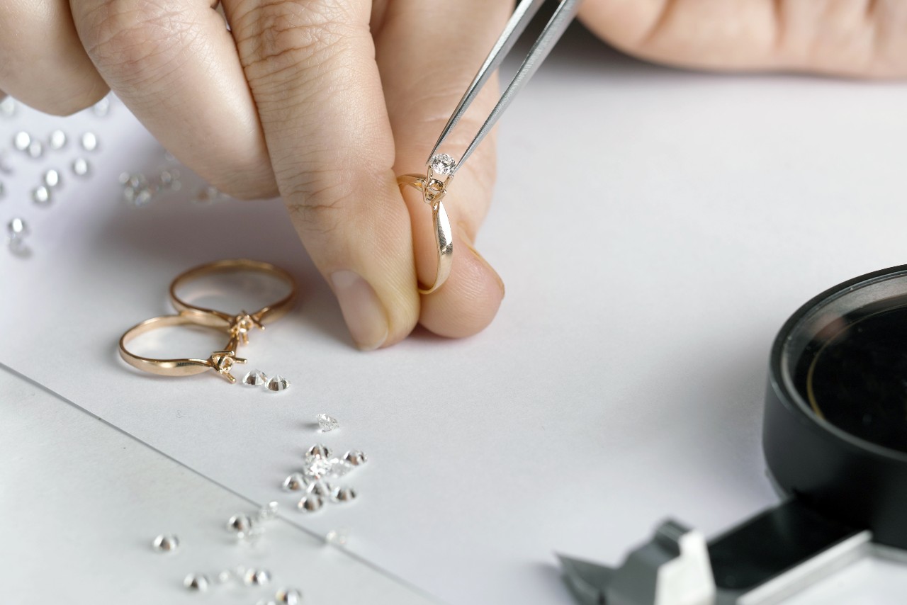 Close up of jeweller’s hand using tweezers to set a diamond on a gold ring, surrounded by loose diamonds and two rings.
