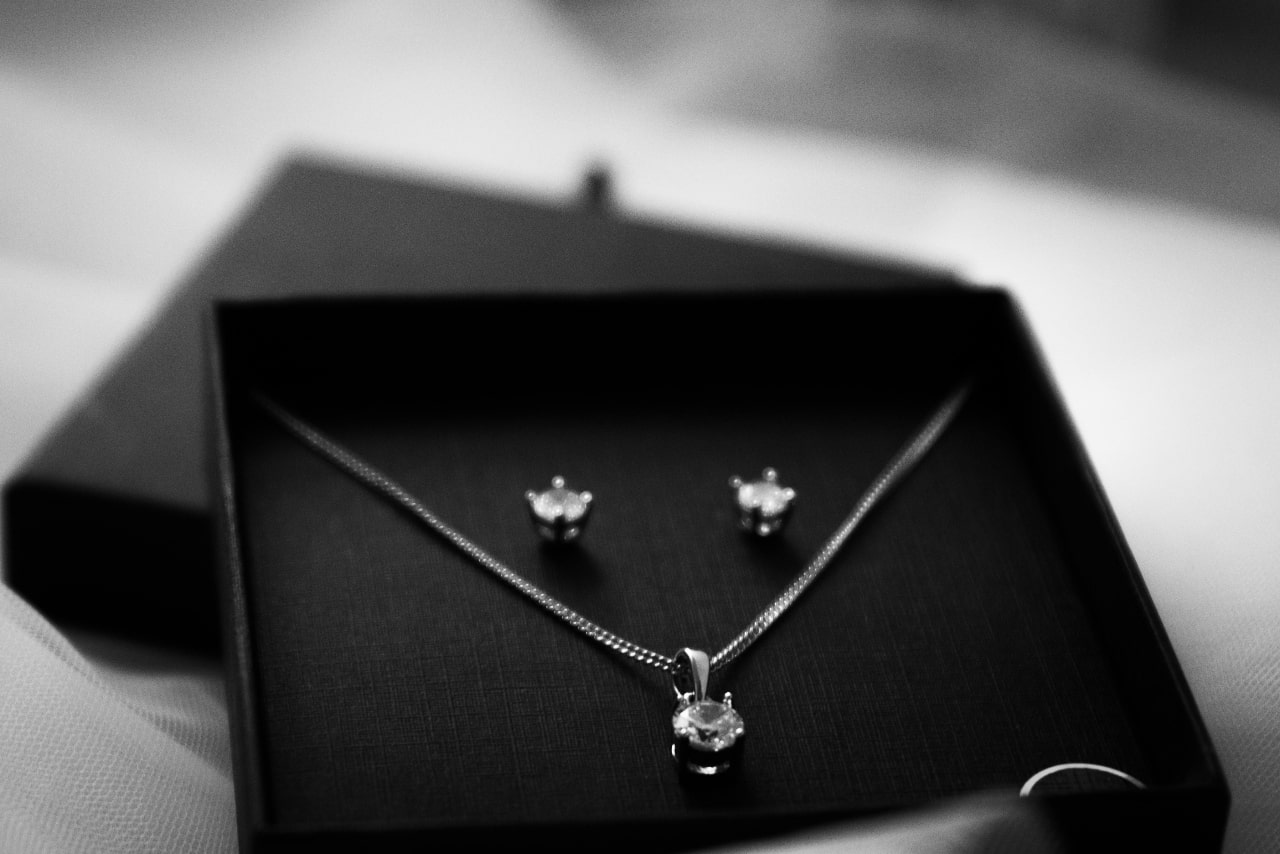 A black gift box displays a silver jewellery set featuring a solitaire pendant necklace and a pair of stud earrings, arranged against a dark fabric background.