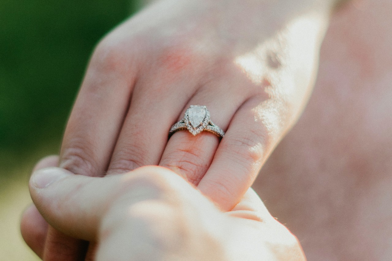 A close-up of a pear-shaped diamond engagement ring with a double halo setting worn on a hand held in natural sunlight.