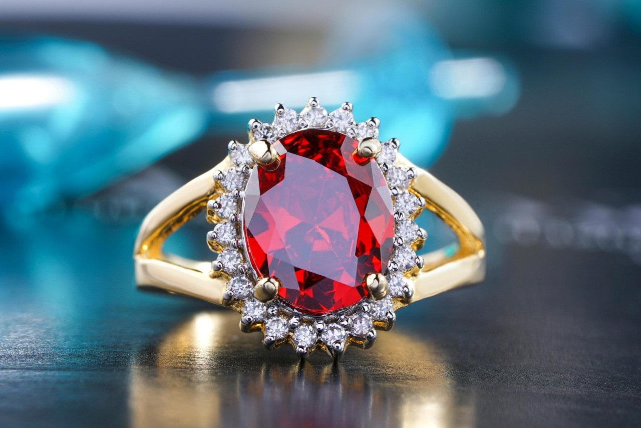 A close-up of a split-shank yellow gold ring featuring an oval-cut red gemstone surrounded by a halo of sparkling white diamonds, set against a blurred blue and black background.