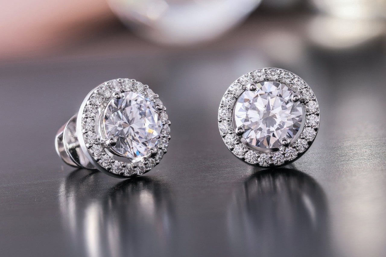 A close up of white gold diamond halo studs against a reflective grey surface.