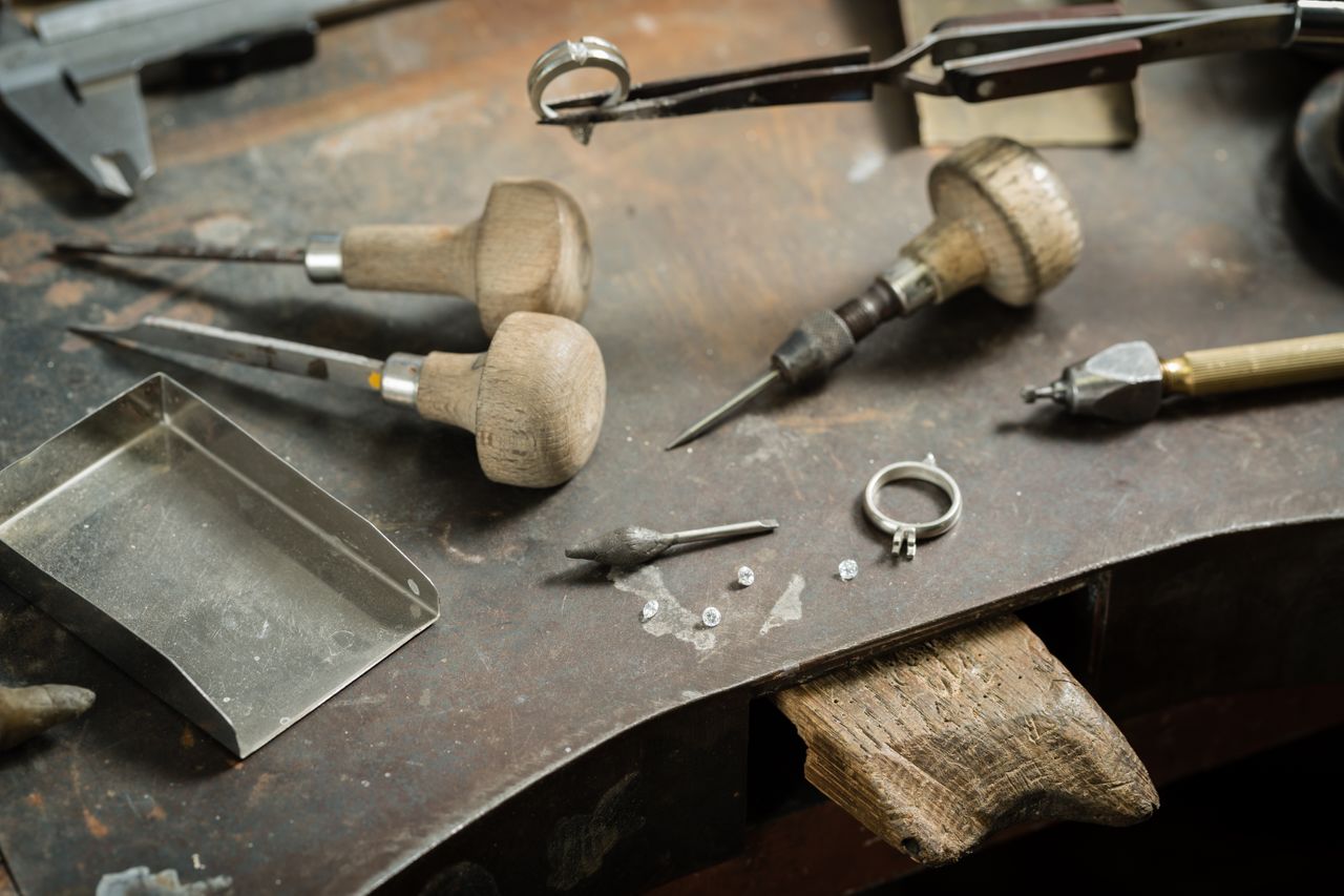 A jeweller's table full of tools, a white gold engagement ring setting, and several diamonds scattered nearby.