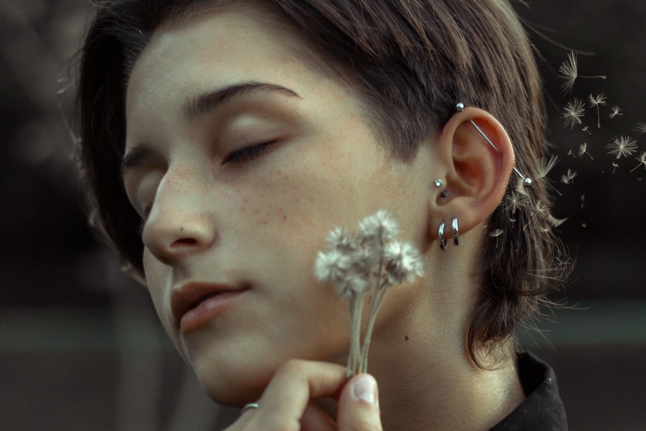 A close-up of a woman with dandelions near her head, showcasing several silver earrings and piercings.