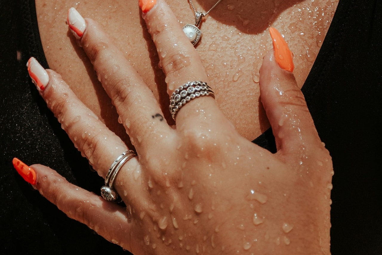 A close-up of a woman’s hand with long manicured nails and silver stackable rings covered with drops of water.